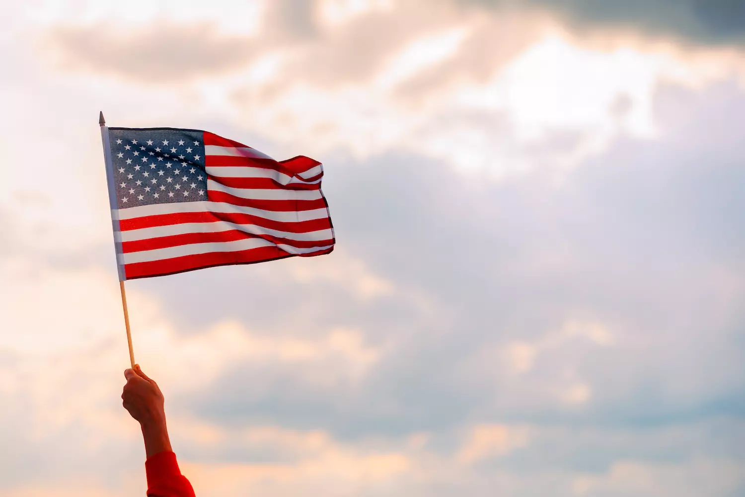 An extended arm waves an American flag in the air in a sunset with some clouds and sun rays in the background. How much patriotic emphasis in worship is too much? How much is too little? Are we mixing two kingdoms?