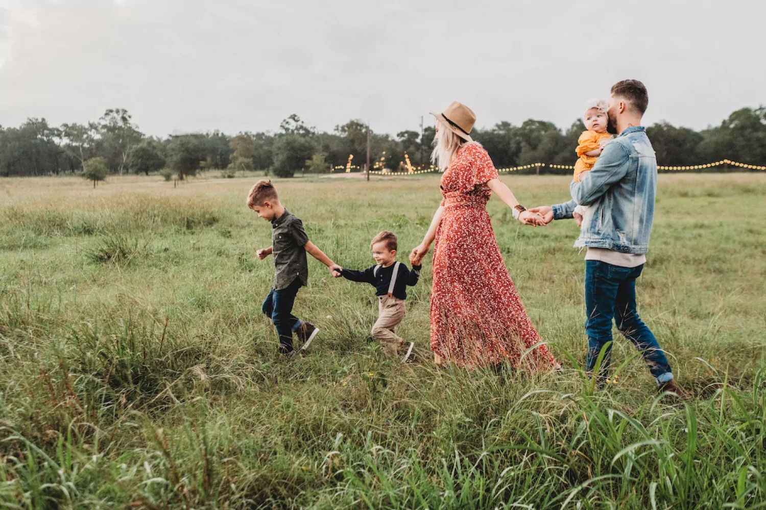 a young family with a mother, father, and three kids walks through a grassy field hand in hand