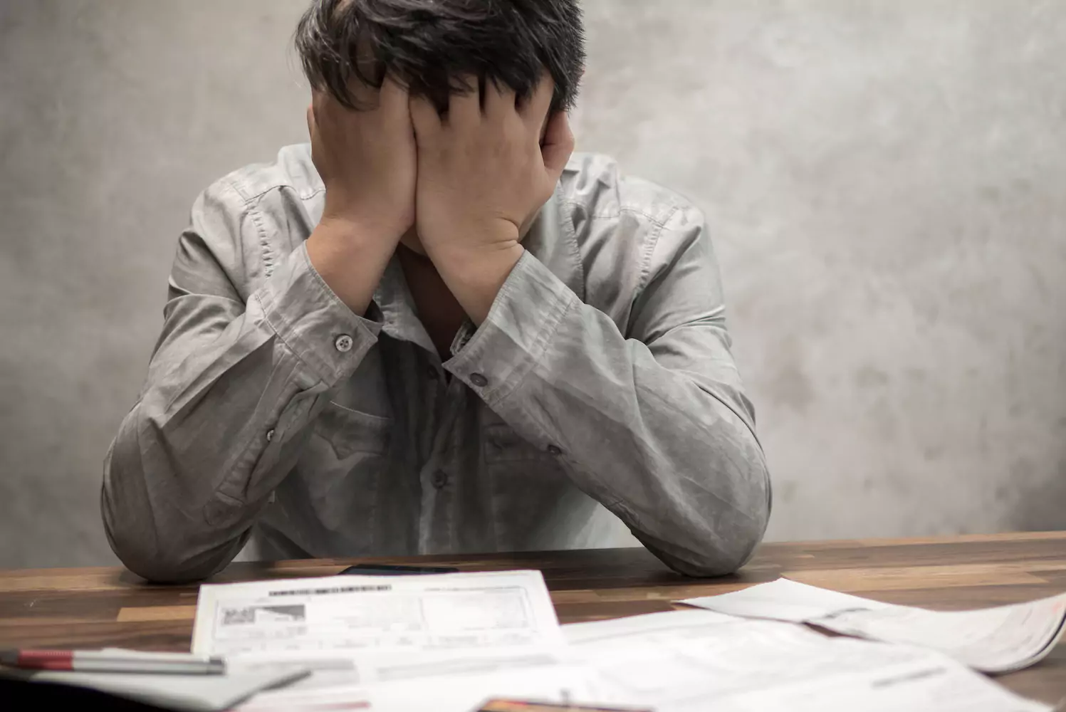 A man sits over a pile of financial papers, stressed out, with his hands covering his face in frustration.