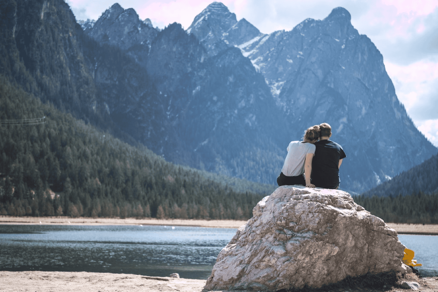 a young wife rests her head on her husbands shoulder as they sit on a rock overlooking a mountain lake