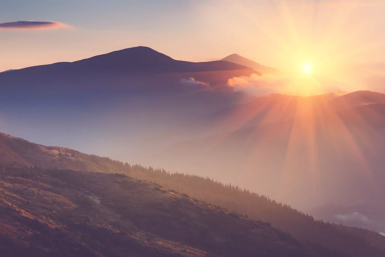 A photo of the sun setting over some mountains. The sun rays break through some haze and illuminate the hillside near the mountain.