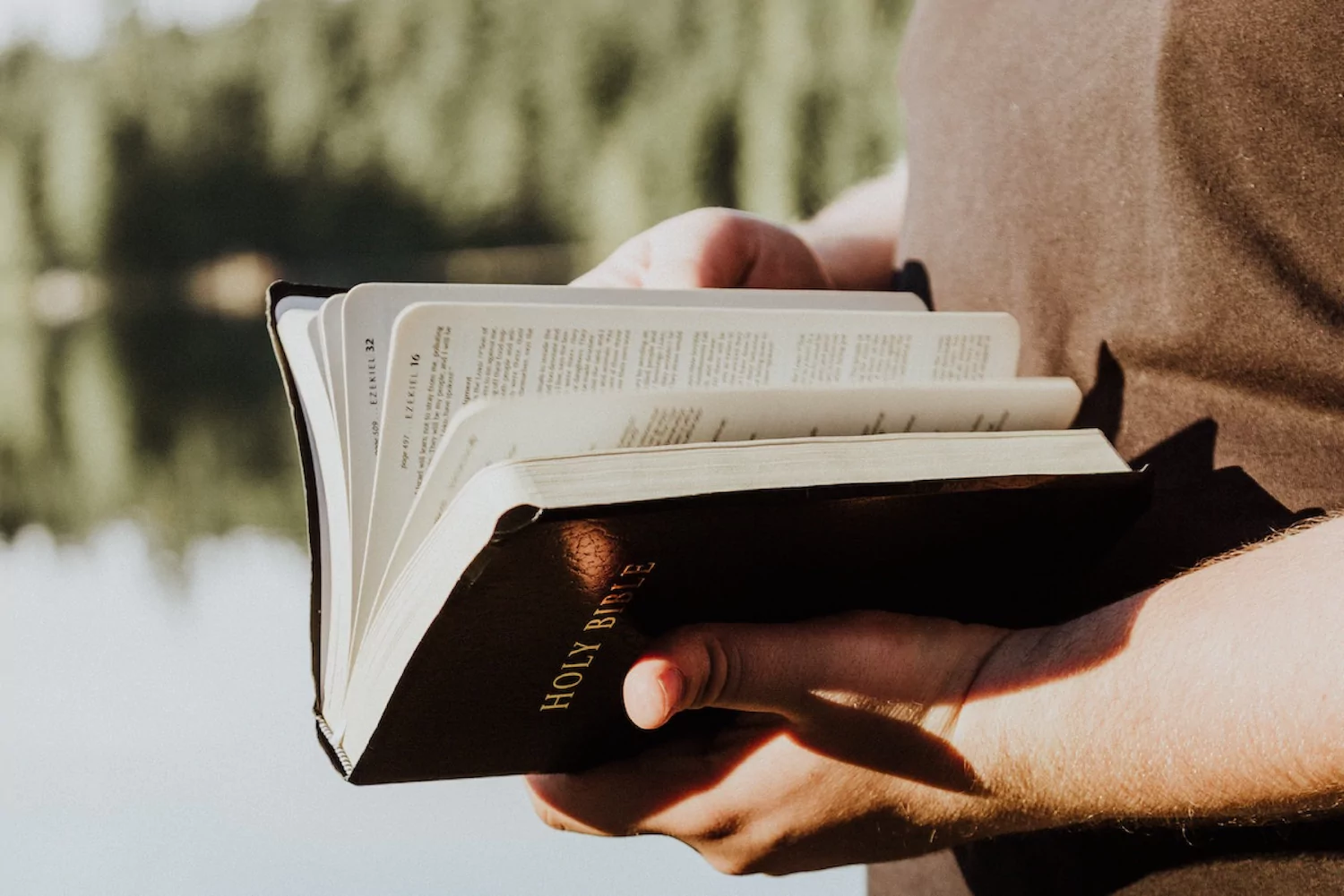 a pastor stands next to a lake with his bible open and pine trees in the background