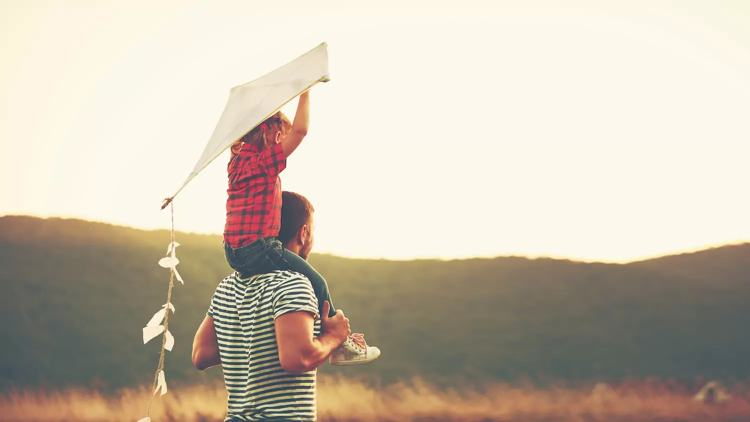 a dad carries his pastor's kid with a kite on his shoulders as they walk through a meadow at golden hour