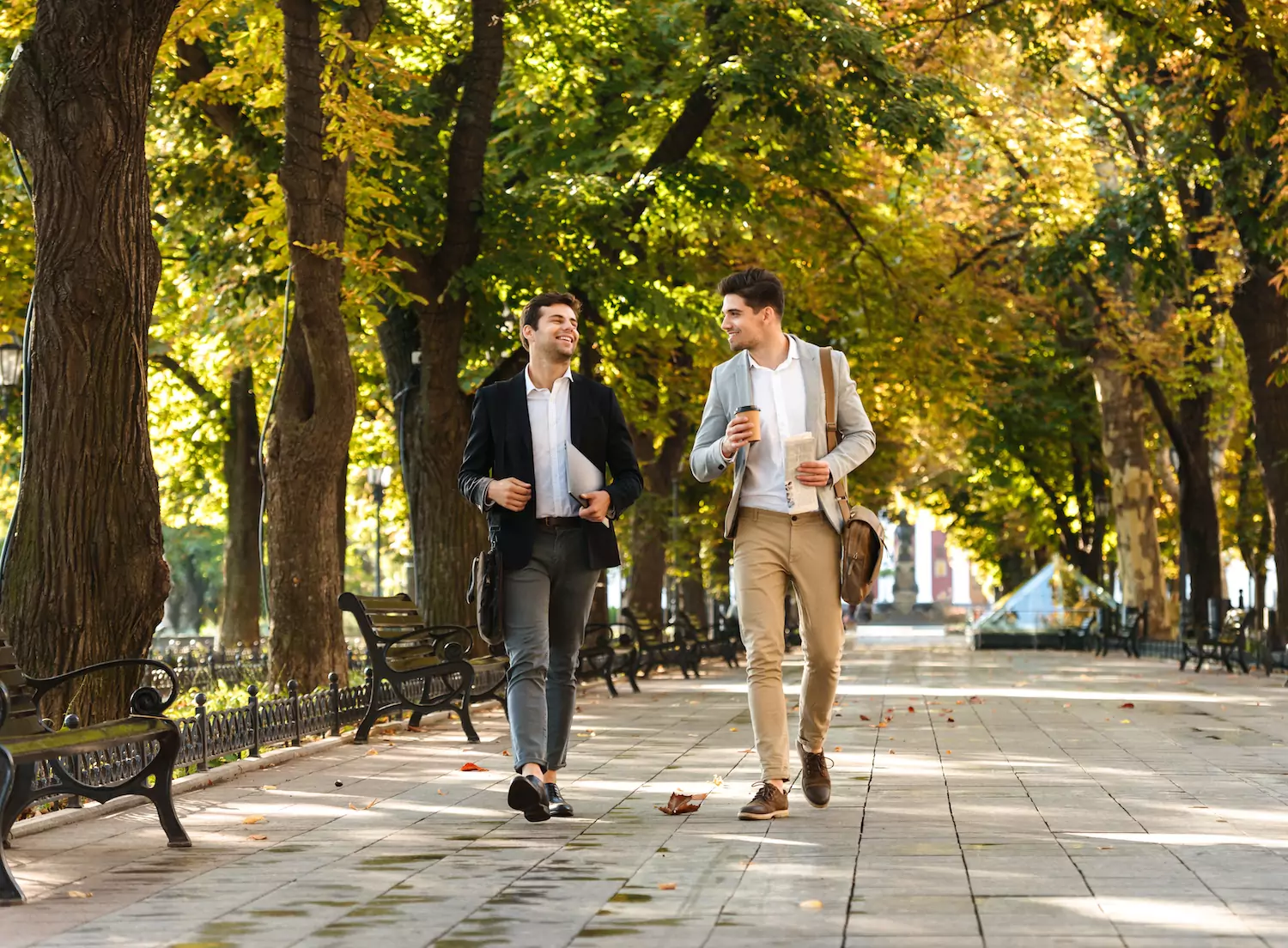 Two pastors walk together as friends, displaying biblical leadership attitudes as they walk down a tree-lined sidewalk at a park in their sport jackets.